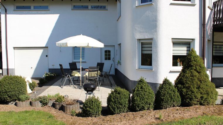 A cozy terrace area with a table and chairs under a sunshade. Some small trees and a barbecue are also visible.