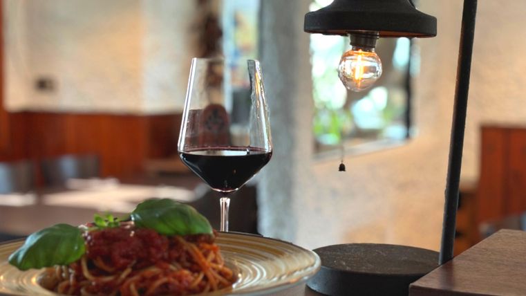 Een bord spaghetti met tomatensaus en verse basilicum. Daarnaast staat een glas rode wijn onder een hangende lamp.