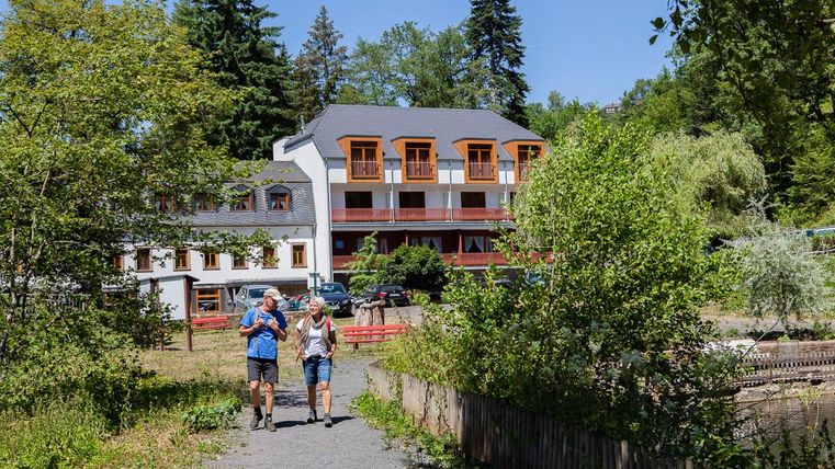 Ein Paar spaziert auf einem Weg, umgeben von Bäumen und Grün. Im Hintergrund ist ein Gebäude mit weißen Wänden und orangefarbenen Fenstern zu sehen.