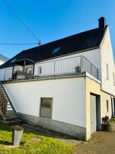 Ein modernes Haus mit einem schrägen Dach und einem Balkon. Die Fassade ist teilweise weiß und es gibt einen kleinen Gartenbereich.