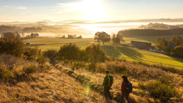 Zwei Wanderer auf einem Pfad in einer hügeligen Landschaft bei Sonnenaufgang.