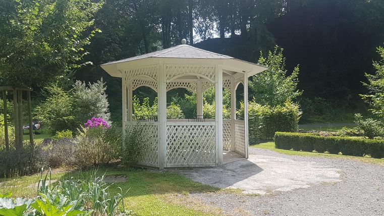 Ein weißer Pavillon steht in einem Garten mit üppigem Grün. Um ihn herum sind Blumen und Sträucher angeordnet.