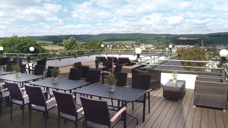 Eine gemütliche Dachterrasse mit modernen Möbeln. Im Hintergrund sind grüne Hügel und ein blauer Himmel mit Wolken zu sehen.