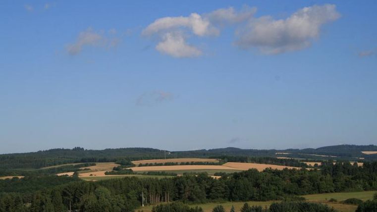 Eine malerische Landschaft mit grünen Wäldern und weiten Feldern. Der Himmel ist klar und die Wolken sind leicht verteilt.