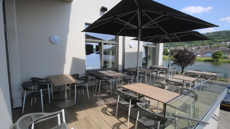 A cozy terrace with tables and chairs is shaded by a large sun umbrella. In the background, a tranquil water landscape can be seen.