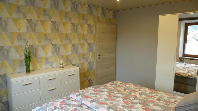 Ein modernes Schlafzimmer mit geometrischem Tapetenmuster und einem gemütlichen Bett. Der Raum ist hell und freundlich, mit einem weißen Schrank und einem Fenster.