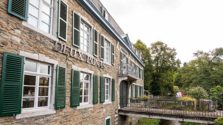 Een historisch gebouw met groene luiken en een rivier op de voorgrond. De omgeving is gekenmerkt door bomen en natuur.