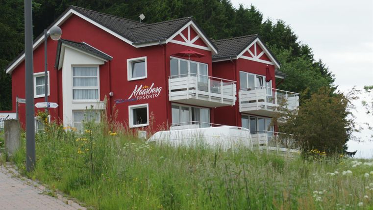 Zwei rot gestrichene Häuser mit Balkonen in einer grünen Umgebung. Im Hintergrund sind Bäume zu sehen und der Himmel ist bewölkt.