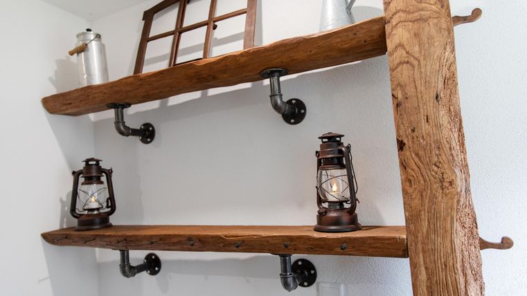 Ein rustikales Regal aus Holz mit zwei Regalböden. Auf den Regalen stehen alte Laternen und an der Wand hängt ein kleines Fenster.