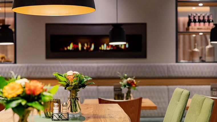 Ein stilvolles Restaurant mit Holztischen und modernen Stühlen. Blumenarrangements und eine gemütliche Beleuchtung schaffen eine einladende Atmosphäre.