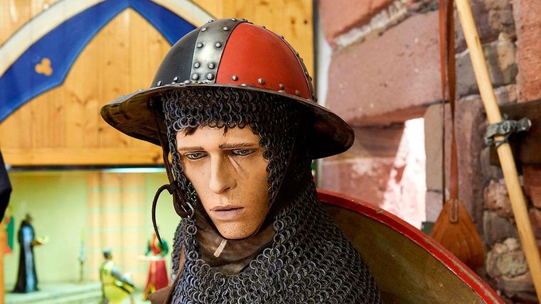 A realistic figure of a knight in chainmail and a striking helmet. In the background, other historical objects are visible.