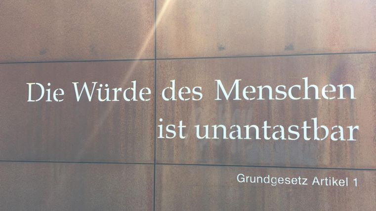 Ein Wandtext, der besagt: „Die Würde des Menschen ist unantastbar. Grundgesetz Artikel 1.“ Der Schriftzug ist auf einer modernen Oberfläche angebracht.