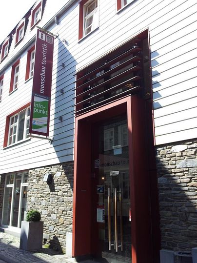 Ein moderner Eingangsbereich mit großen Glastüren. Das Gebäude kombiniert Holz und Stein und enthält ein Schild für einen Infopunkt.