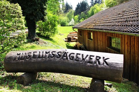 A wooden sign with the engraving "Museum Sawmill" stands in front of a rustic building. In the background, green meadows and trees are visible.