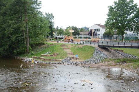 Een rustige rivier met kiezelstenen en een speeltuin op de achtergrond. Omgeven door bomen en een groene weide.