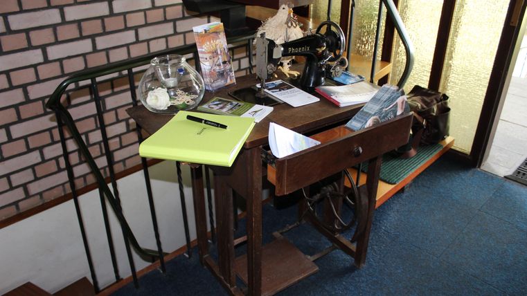 Ein antiker Nähmaschine steht auf einem kleinen Tisch im Flur. Daneben liegen einige Bücher und eine grüne Notizmappe.