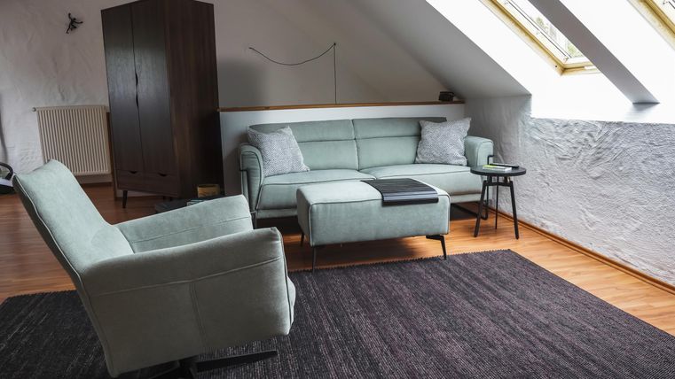 Ein gemütliches Wohnzimmer mit einem hellgrünen Sofa und einem Sessel. Große Fenster sorgen für viel Tageslicht und eine einladende Atmosphäre.