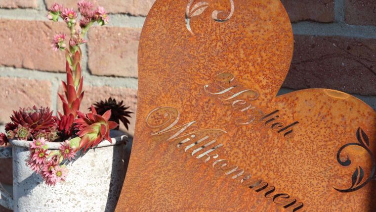 Ein herzförmiges Willkommensschild aus Metall steht neben einem Topf mit bunten Pflanzen. Die warme Farbe des Schildes harmoniert gut mit der grünen und roten Bepflanzung.