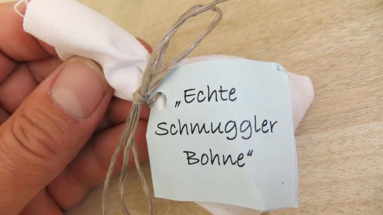 A hand holds a small package with a sticker that says "Real Smuggler Bean." The package is tied with a piece of string and looks like a gift.