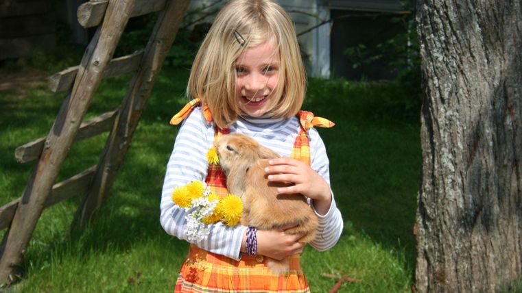 Ein lächelndes Mädchen hält ein Kaninchen in den Armen und trägt ein farbenfrohes Kleid. Im Hintergrund sind grüne Wiese und ein Baum sichtbar.