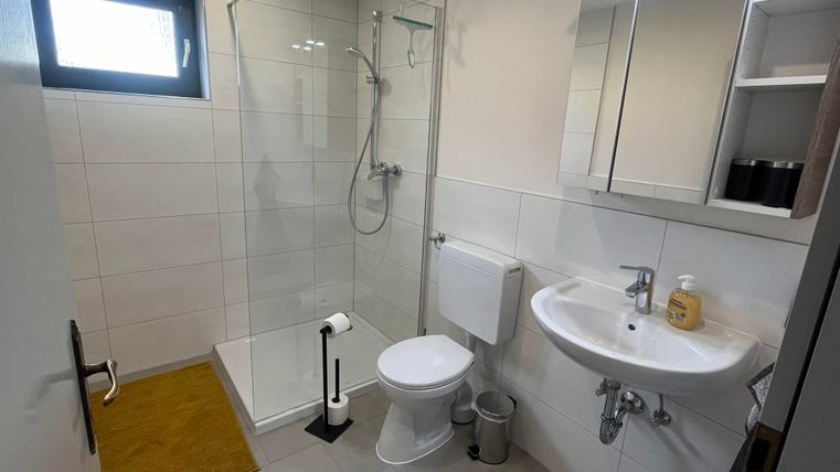 A modern bathroom with a shower cabin and a sink. The walls are light tiled, and there is a small bath mat in a warm color.