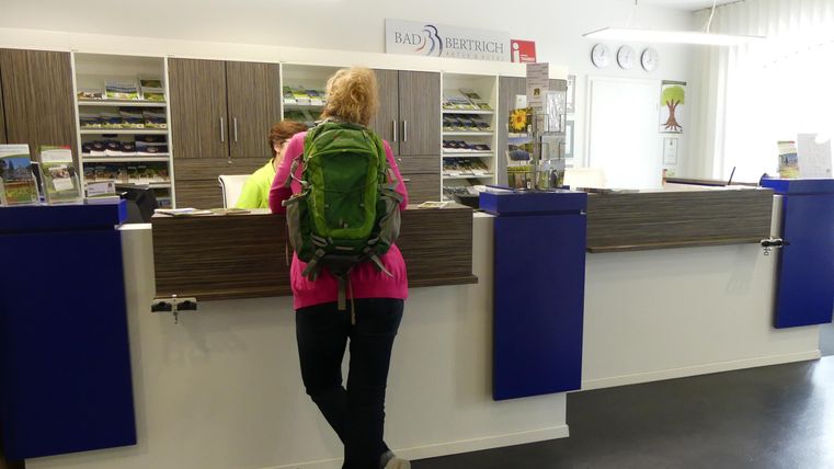 Een persoon met een groene rugzak staat bij een receptiebalie. Het gebied lijkt helder en modern, met planken op de achtergrond.