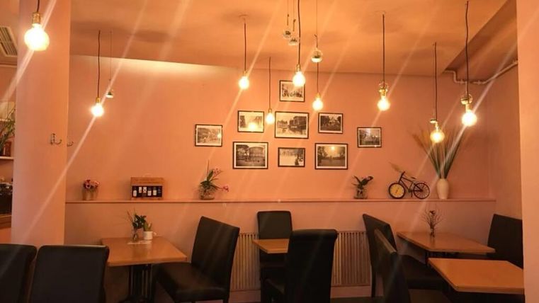 Een sfeervol café met perzikkleurige muren en warme hanglampen. Aan de muren hangen schilderijen, en de tafels en stoelen zijn eenvoudig gerangschikt.