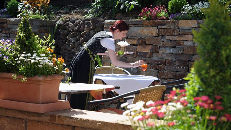 Eine Frau deckt einen Tisch im Freien. Umgeben von bunten Blumen und einer schönen Steinmauer sieht es einladend aus.