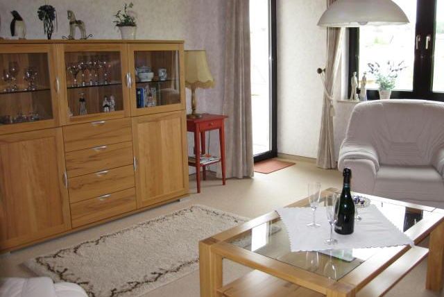 A cozy living room with a wooden cabinet, a modern couch, and a glass table. On the table, there are a bottle of champagne and two glasses, while light streams through large windows.