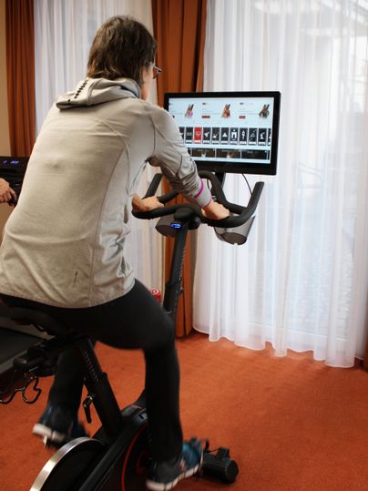 Eine Person trainiert auf einem Indoor-Bike vor einem Bildschirm. Der Bildschirm zeigt eine Auswahl an Fahrradausrüstung.