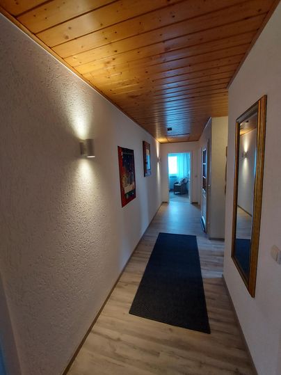 A beautiful hallway with a wooden ceiling and bright walls. At the end of the hallway, there is a door that leads to a bright room.
