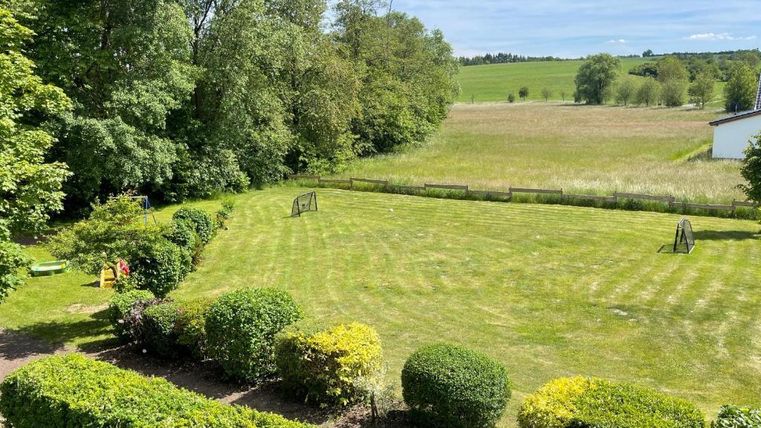 A green meadow surrounded by trees and bushes. In the background, fields can be seen and a blue sky.