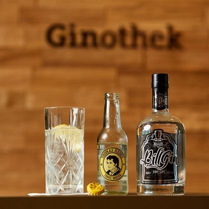 Ein elegantes Glas mit einem Drink, garniert mit einer Zitronenscheibe, steht neben einer Flasche Thomas Henry Tonic Water und einer Flasche BillGin. Im Hintergrund ist das Wort „Ginothek“ zu sehen.