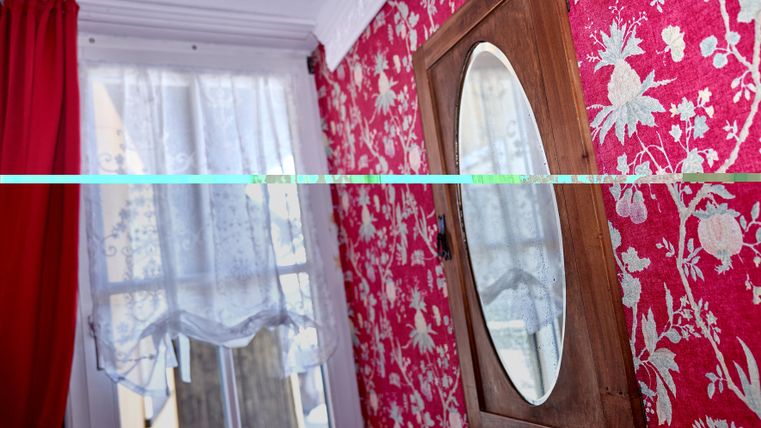 A room with red patterned walls and a wooden mirror. A white curtain hangs in front of the window.