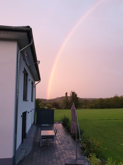 Ein modernes Haus mit einer Terrasse und einem Tisch steht im Vordergrund. Im Hintergrund ist ein bunter Regenbogen über einer grünen Wiese zu sehen.