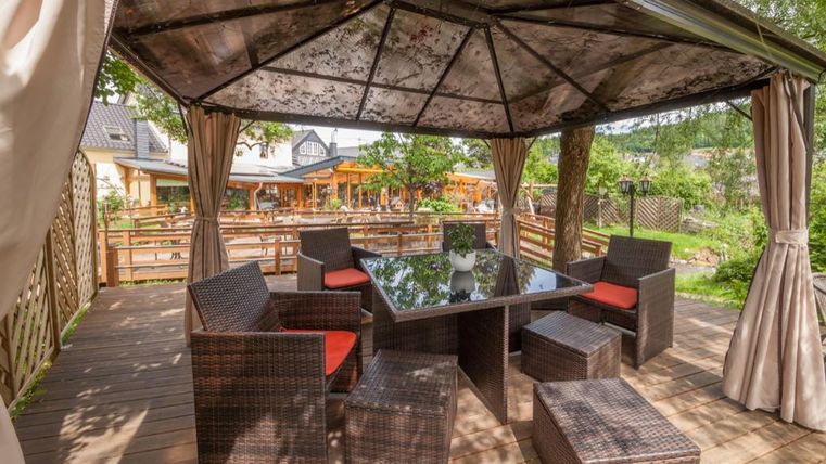 Eine gemütliche Terrasse mit einem Pavillon und Gartenmöbeln. Im Hintergrund sind grüne Pflanzen und eine einladende Atmosphäre zu sehen.