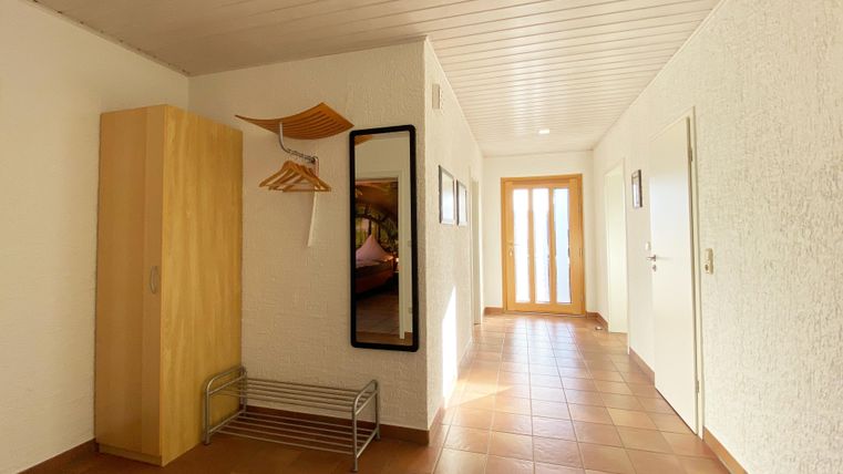 Ein heller Flur mit weißen Wänden und braunen Fliesen. Es gibt einen Holzschrank, einen Spiegel und eine Glasschiebetür, die zu einem weiteren Raum führt.