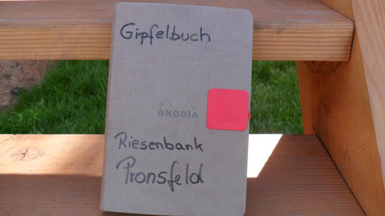 A summit book titled "Riesenbank Pronsfeld" lies on a wooden bench. It has a gray cover and a red label.
