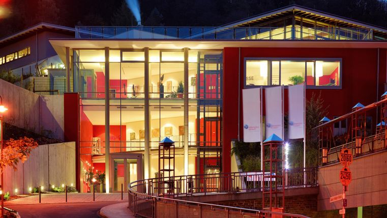 Een modern gebouw met grote ramen en een rode gevel. Het is 's avonds verlicht en omgeven door een zachte landschap.