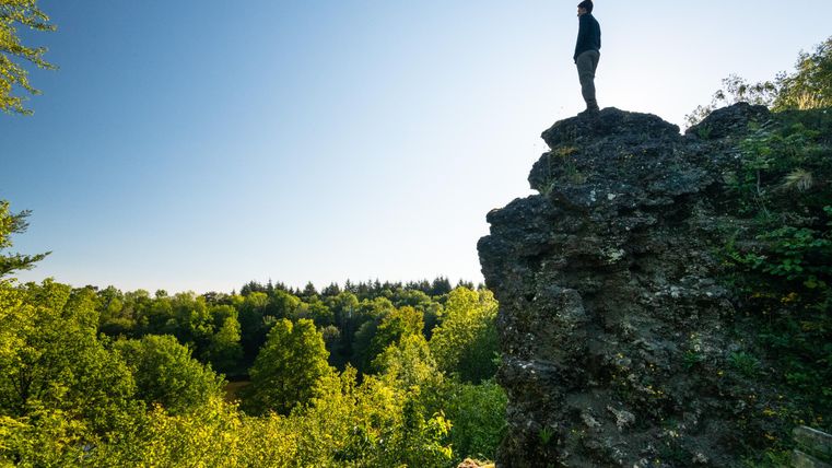 Ein Mann steht auf einem hohen Felsen und blickt auf eine grüne Landschaft. Die Sonne scheint und der Himmel ist klar.