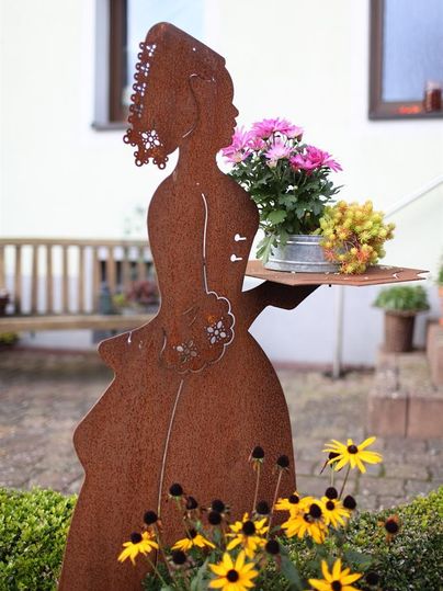 Een decoratieve metalen figuur die een vrouw met bloemen presenteert. Op de achtergrond zijn kleurrijke bloemen en een houten bank te zien.