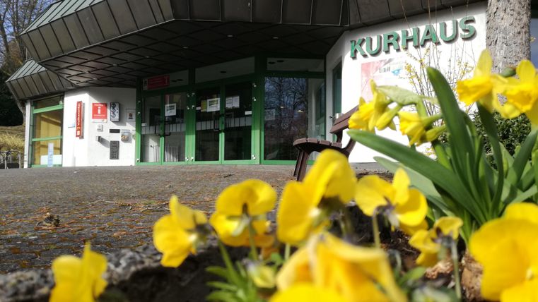 Das Bild zeigt den Eingangsbereich eines Kurhauses mit großen Fenstern und einem modernen Dach. Im Vordergrund blühen gelbe Blumen, die die Szene lebendig machen.