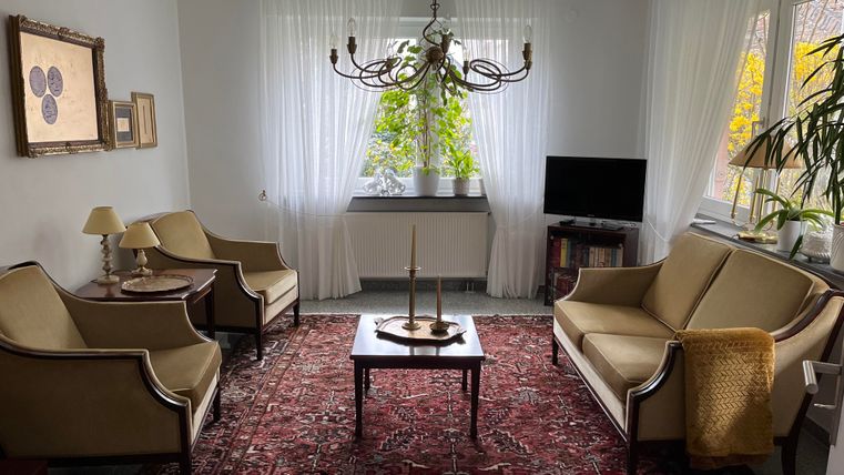 Ein gemütliches Wohnzimmer mit zwei beigen Sesseln und einer Couch. In der Mitte steht ein Tisch und die Fenster sind mit weißen Vorhängen dekoriert.