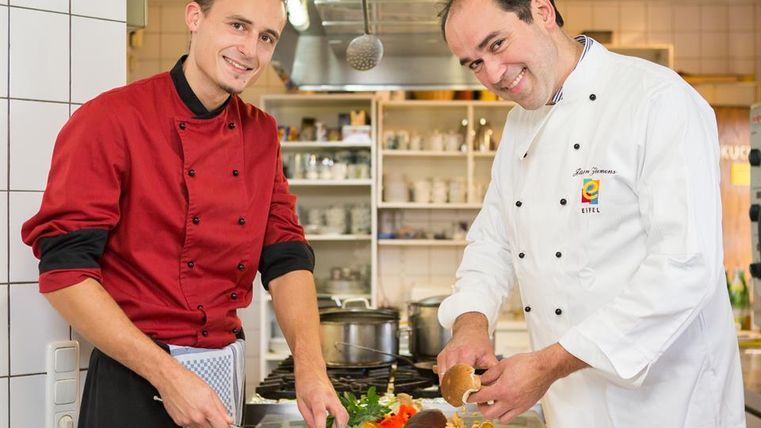 Zwei Köche arbeiten in einer modernen Küche. Der eine schneidet Fisch, während der andere Gemüse vorbereitet.