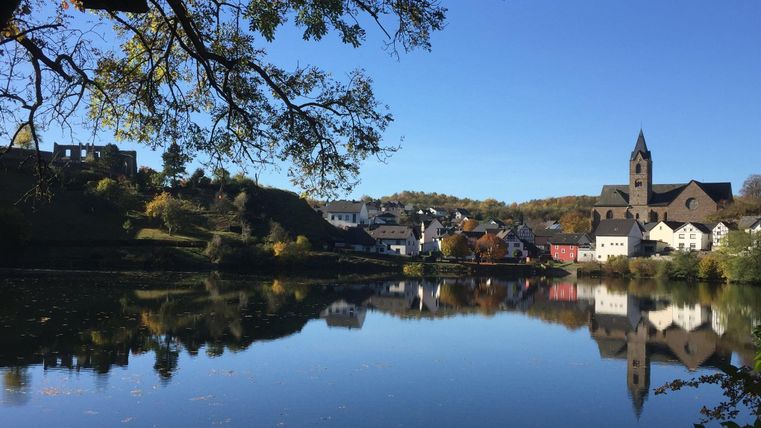 A picturesque village by the river with a clear reflection in the water. In the background, houses and a church tower can be seen, surrounded by trees and blue sky.