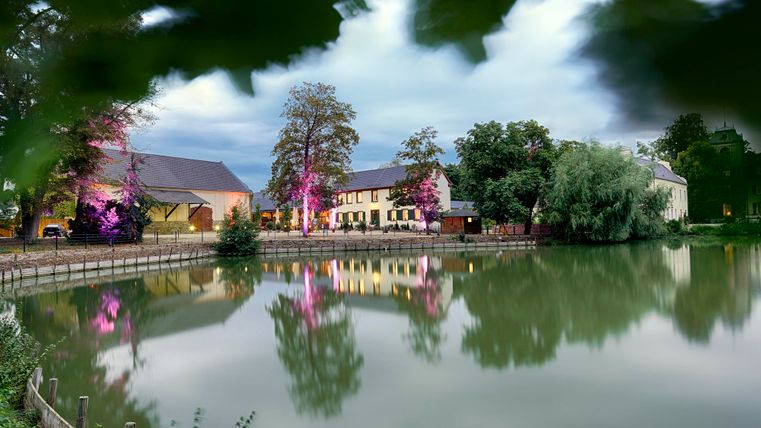Een rustige meer met reflecterende gebouwen en bomen op de achtergrond. De scène is omgeven door zacht licht en straalt een vredige sfeer uit.