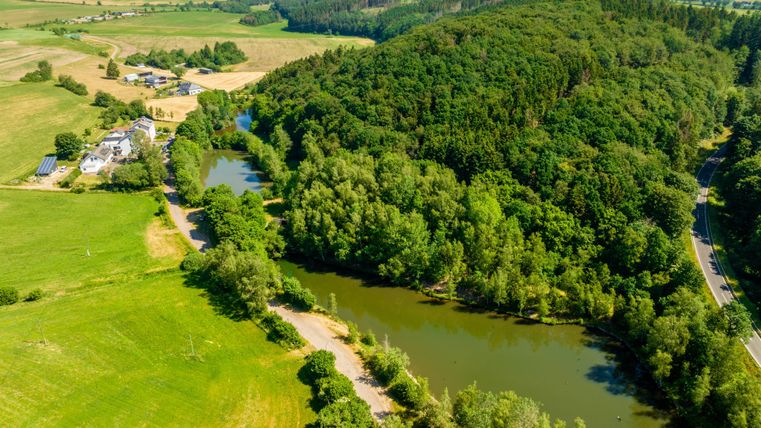A picturesque landscape with a river, surrounded by lush greenery and wooded hills. On the left side, fields and a building are visible.