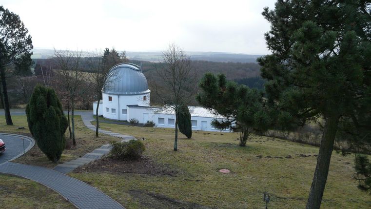 Eine Sternwarte steht in einer grünen Landschaft mit Bäumen. Der Himmel ist bewölkt und die Umgebung wirkt ruhig und idyllisch.
