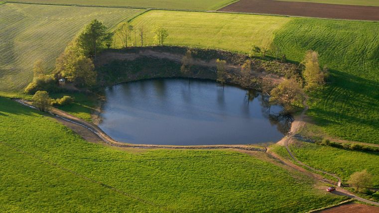 Ein ruhiger Teich umgeben von grünen Feldern und vereinzelten Bäumen. Die Landschaft ist weitläufig und freundlich gehalten.