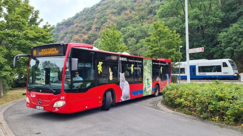 A red bus is rounding a curve while a train stops nearby. In the background, green trees and a mountainous slope can be seen.